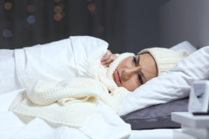 Woman wearing winter hat and scarf scowls and covers up in bed.