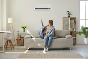 woman sits on sofa and adjusts temperature of ductless heating unit