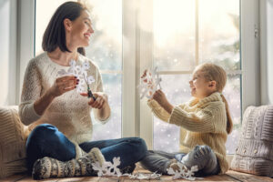 woman and child sit in front of window making snowflakes in winter